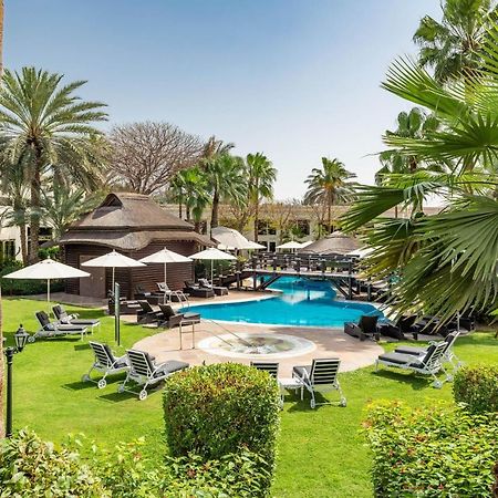 Le Meridien Dubai Hotel & Conference Centre Екстериор снимка The photo shows a serene outdoor scene featuring a swimming pool surrounded by lush greenery. There are several palm trees and shrubs providing a tropical atmosphere. Lounge chairs are arranged around the pool, along with shaded areas created by umbr