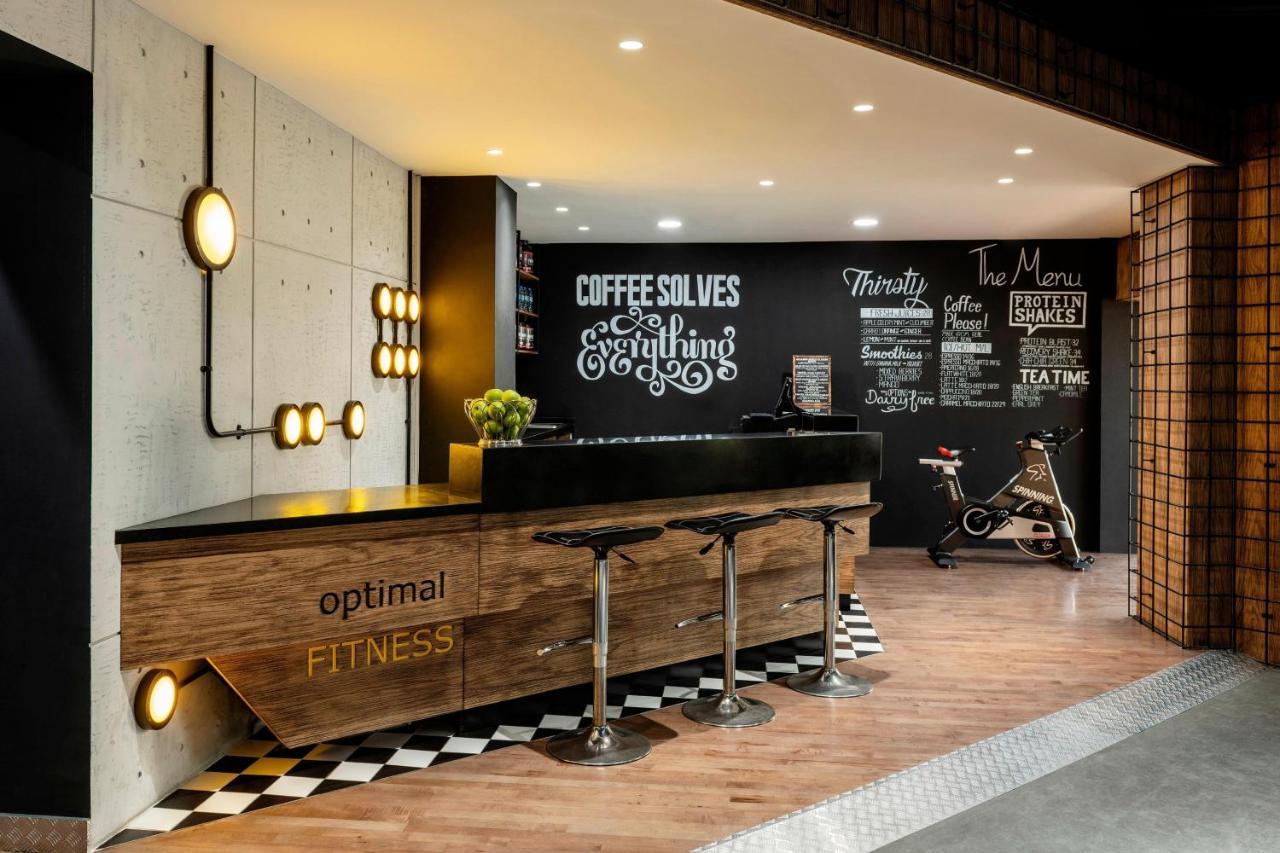Le Meridien Dubai Hotel & Conference Centre Екстериор снимка The photo showcases a modern fitness reception area. A sleek wooden reception desk with the words "optimal FITNESS" is positioned prominently. Behind the desk, a black wall features motivational phrases and chalkboard-style text, including "COFFEE SO