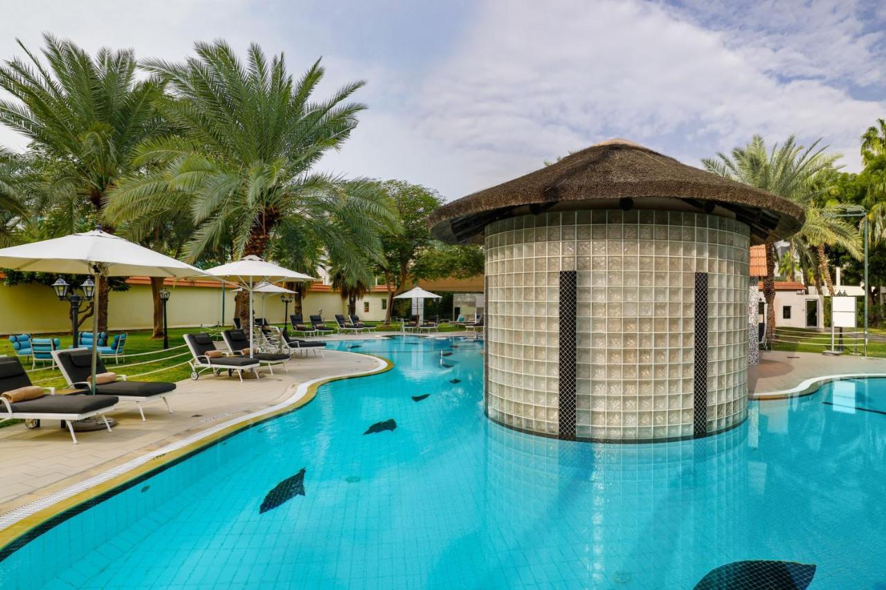 Le Meridien Dubai Hotel & Conference Centre Екстериор снимка The photo shows a luxurious pool area surrounded by palm trees. The pool has a unique design with a circular, glass-brick structure in the center, which may serve as a bar or lounge. There are lounge chairs placed around the pool, and several white u