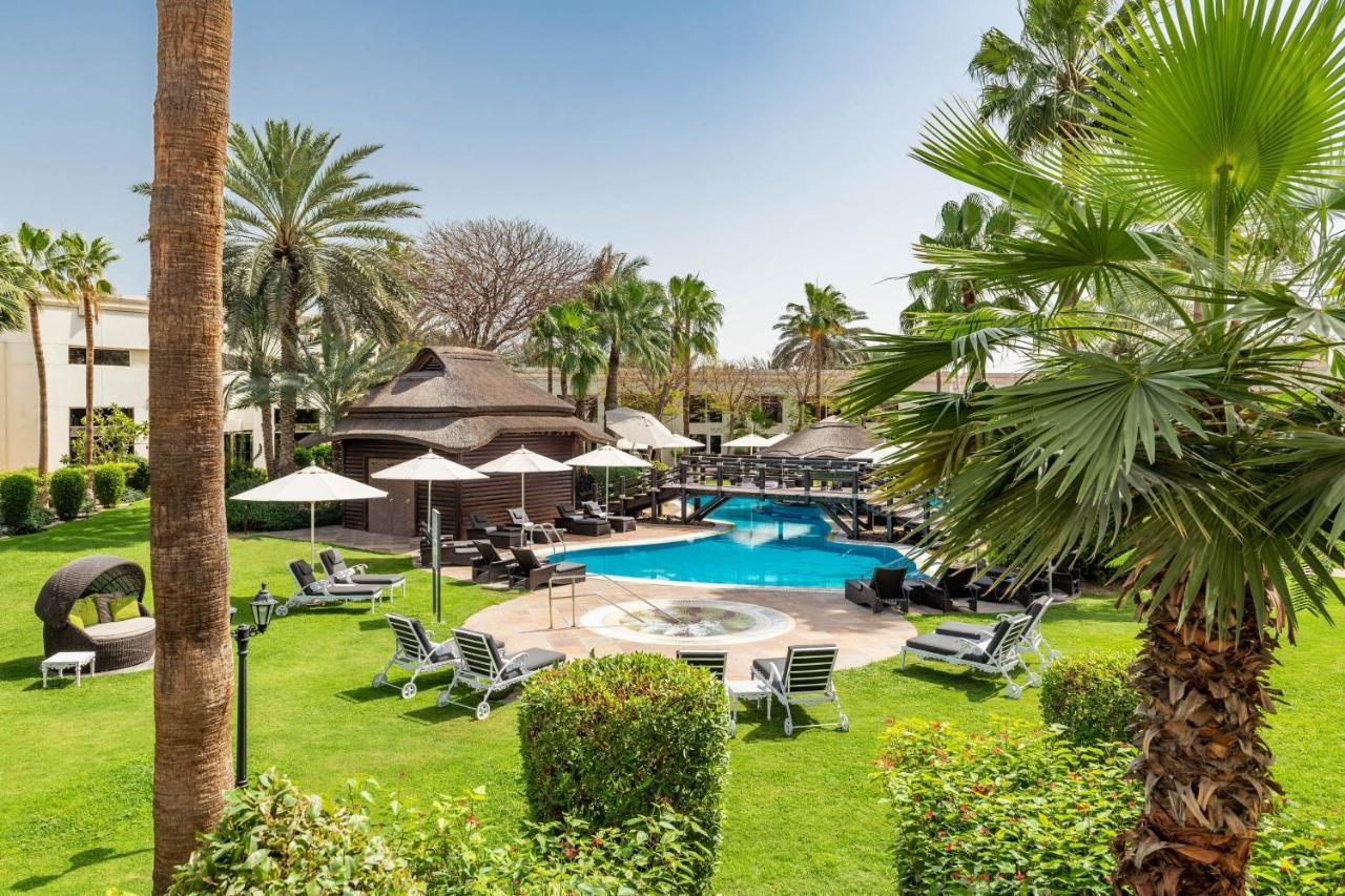 Le Meridien Dubai Hotel & Conference Centre Екстериор снимка The photo shows a serene outdoor scene featuring a swimming pool surrounded by lush greenery. There are several palm trees and shrubs providing a tropical atmosphere. Lounge chairs are arranged around the pool, along with shaded areas created by umbr