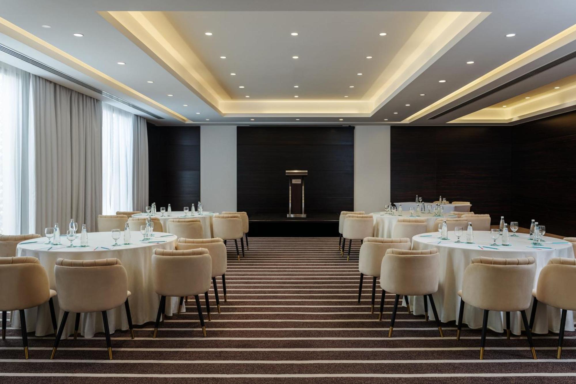 Le Meridien Dubai Hotel & Conference Centre Екстериор снимка The photo shows an elegantly designed conference or banquet room. There are round tables covered with white tablecloths, each accompanied by plush, light-colored chairs. The flooring features a striped pattern. The room is well-lit with recessed ceil